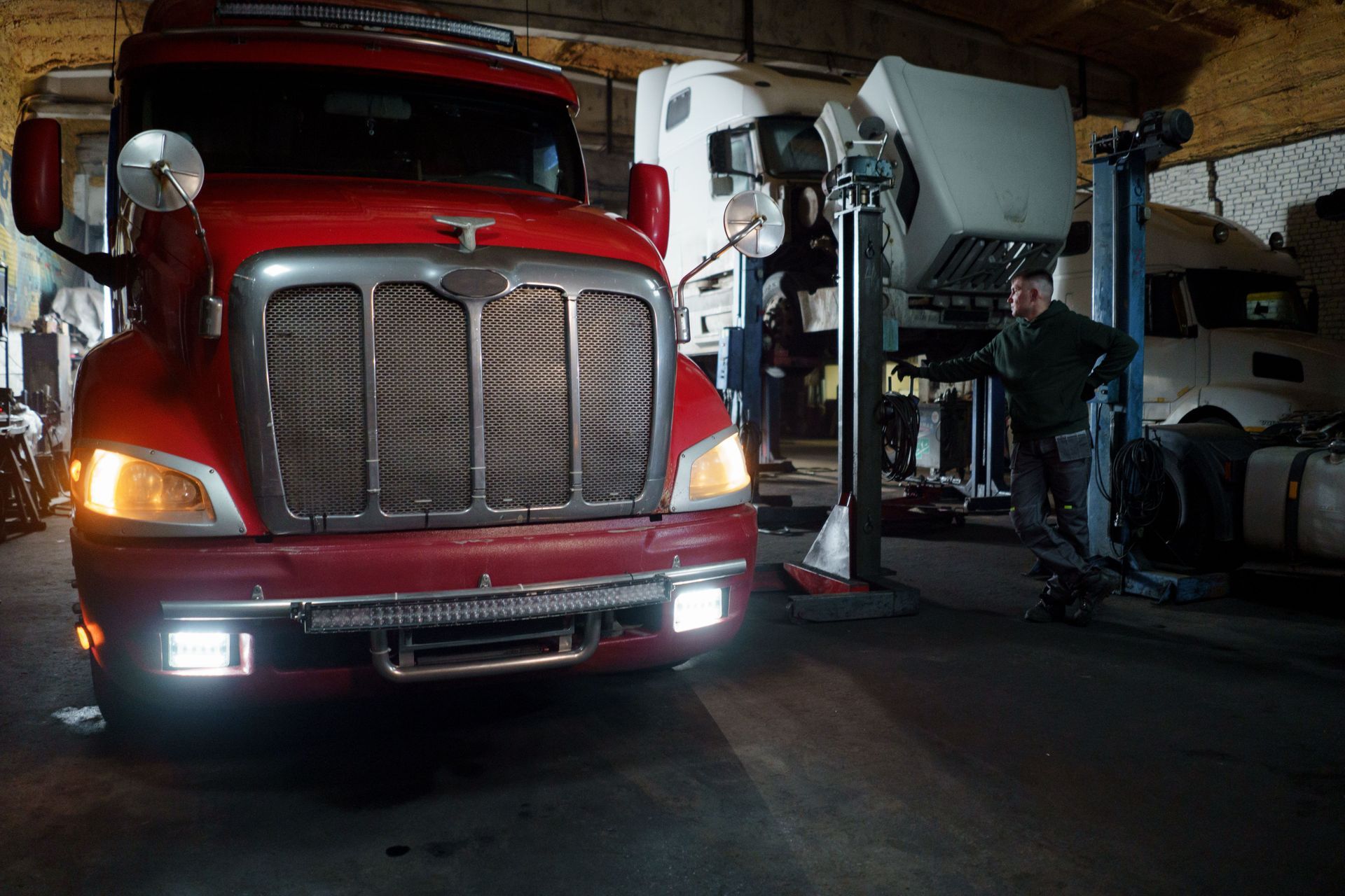 Red semi-truck in a garage beside a white cement mixer truck, with headlights on
