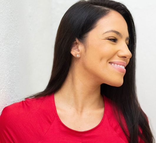 Iris with dark hair smiles, wearing a red shirt, against a white wall.