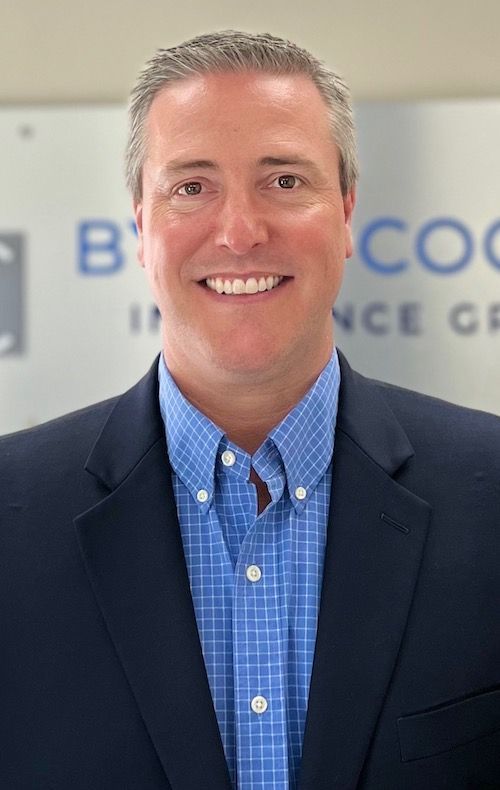 A man in a suit and blue plaid shirt is smiling for the camera.