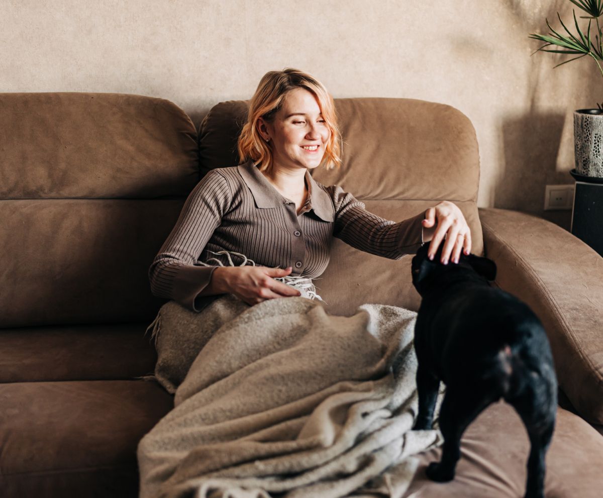 A woman is sitting on a couch petting a black dog.