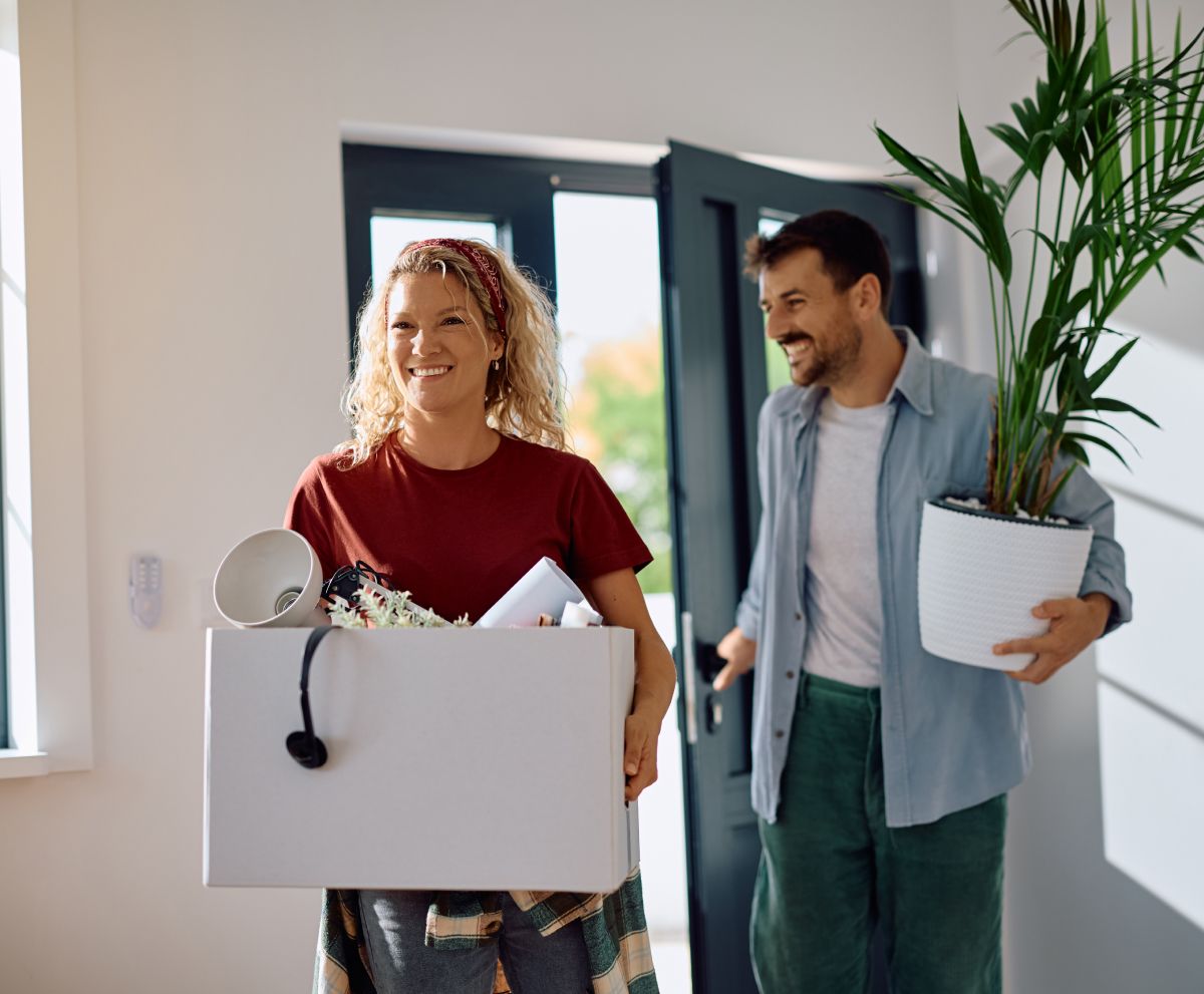 A man and a woman are carrying boxes and potted plants into their new home.