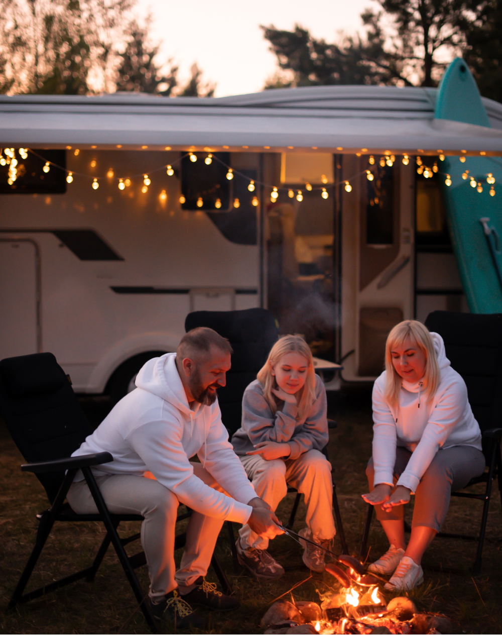 A group of people are sitting around a campfire in front of a trailer.