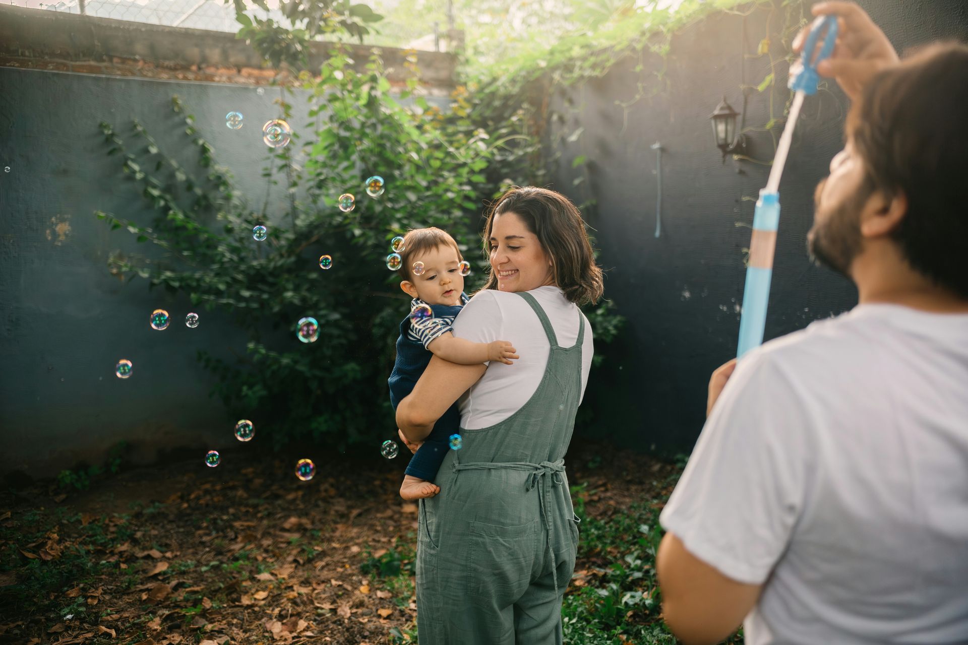 A woman is holding a baby while a man blows soap bubbles.