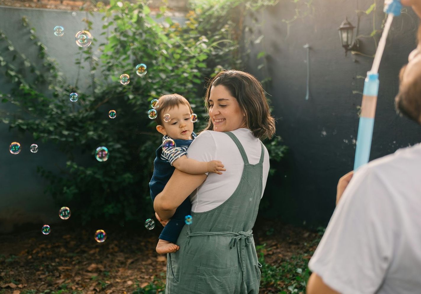 A woman is holding a baby while a man blows soap bubbles.