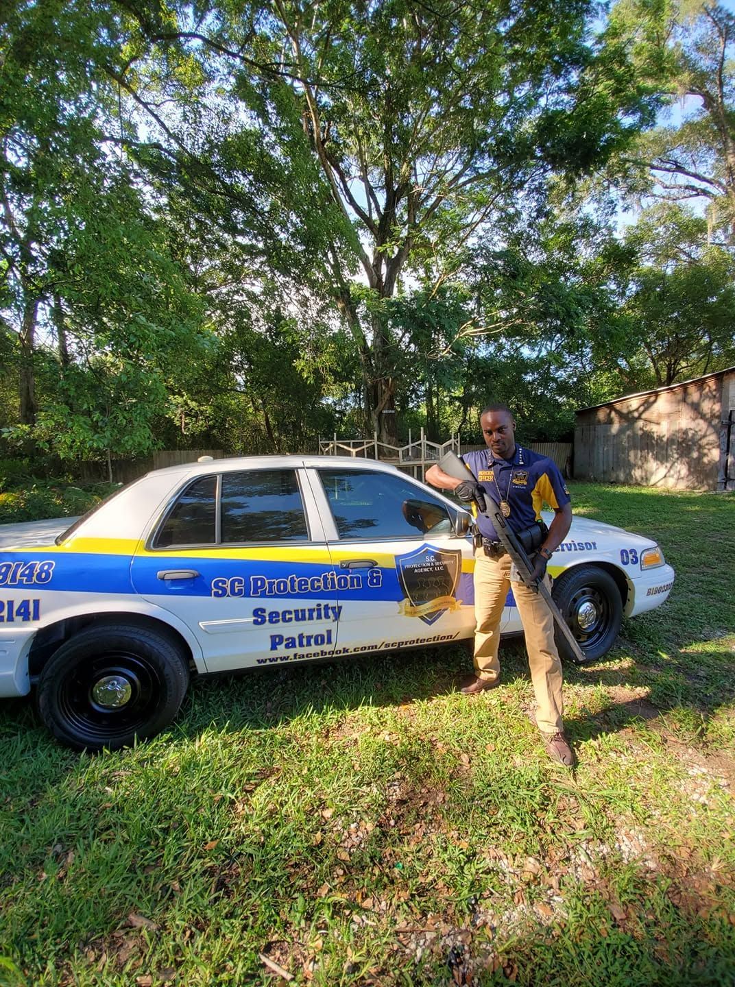 Police car parked in grass with an officer standing beside it under trees