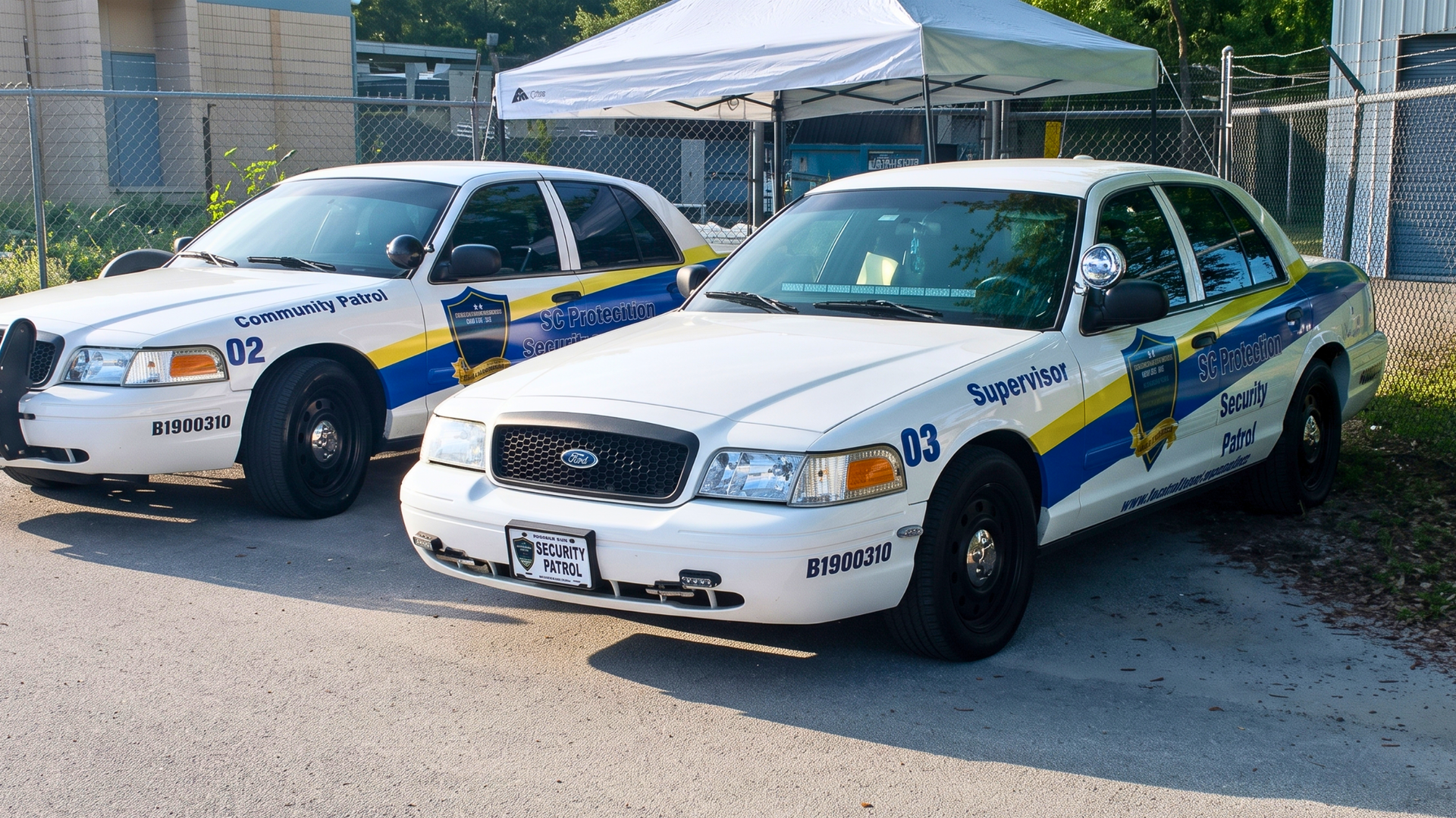 Two parked white police cars with blue-and-yellow markings under a canopy on a sunny street.