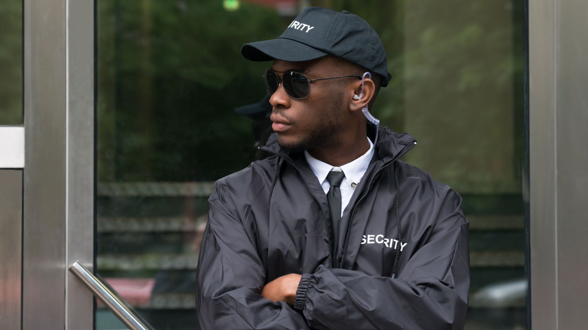 A uniformed security guard in a black cap and sunglasses stands with arms crossed outside a glass building.