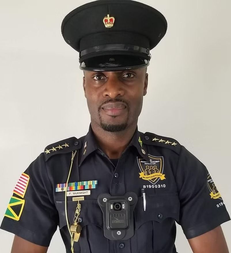 Uniformed security officer standing against a plain wall, wearing a black cap and body camera.