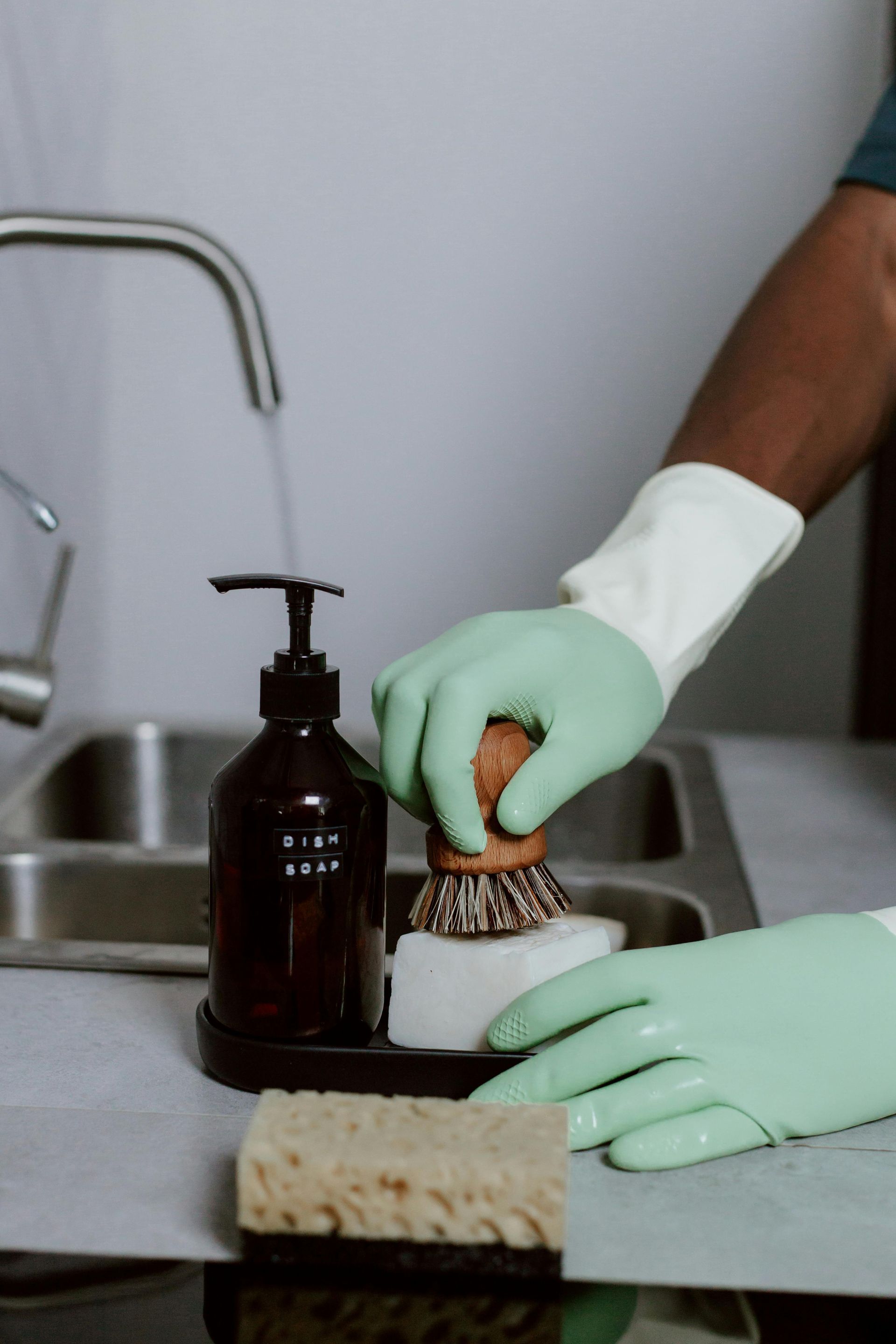 Gloved hands scrubbing a dish at a kitchen sink beside a soap dispenser and sponge.