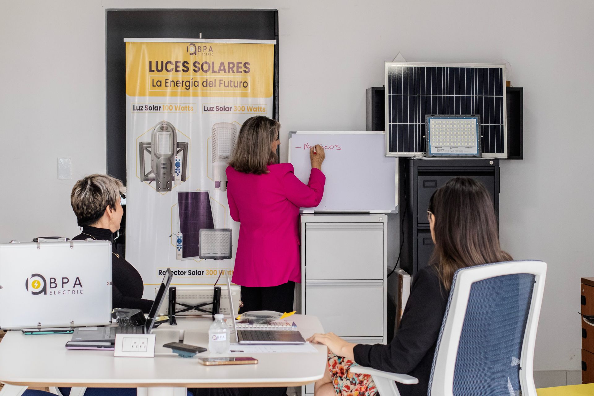 Two people in an office examining a wall-mounted solar panel display with a “LUCES SOLARES” banner.