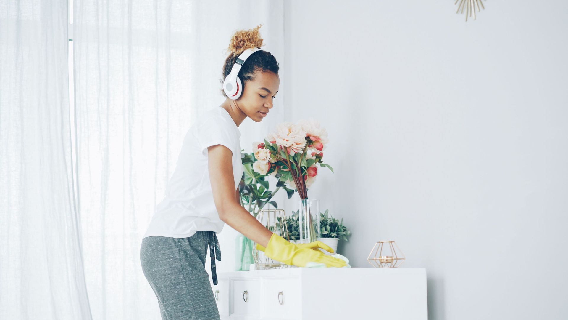 Woman in headphones wiping a white dresser beside flowers in a bright room
