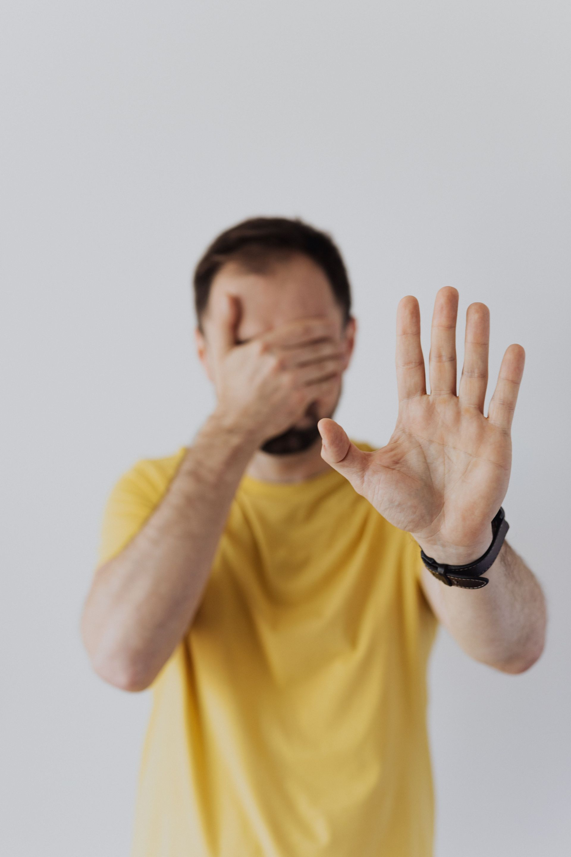 Person in a yellow shirt holding one hand up to the camera while covering their face with the other