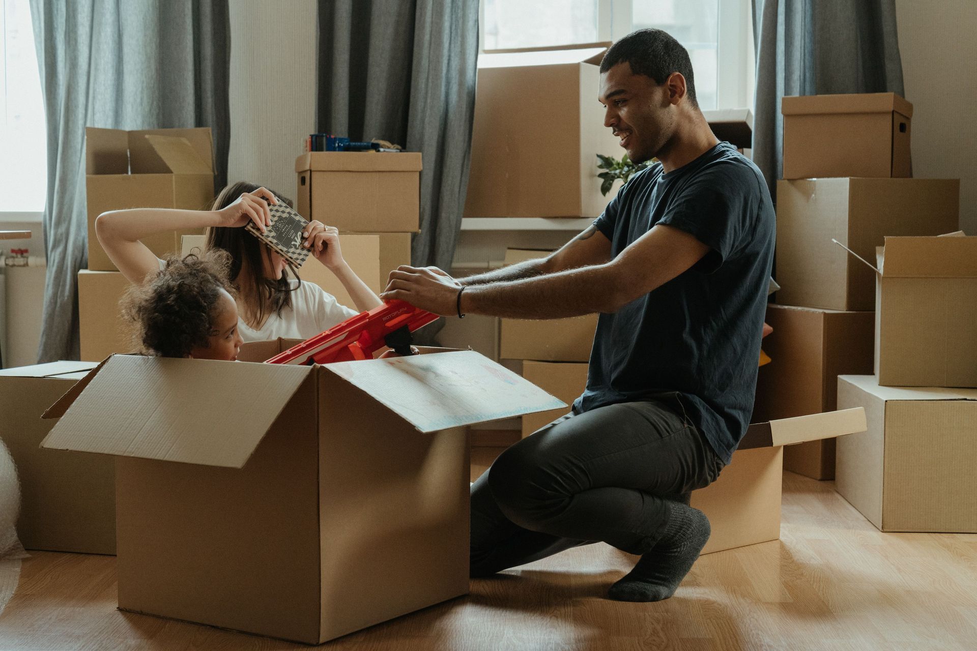 Man unpacking a moving box while a child reaches out, surrounded by stacked boxes in a living room
