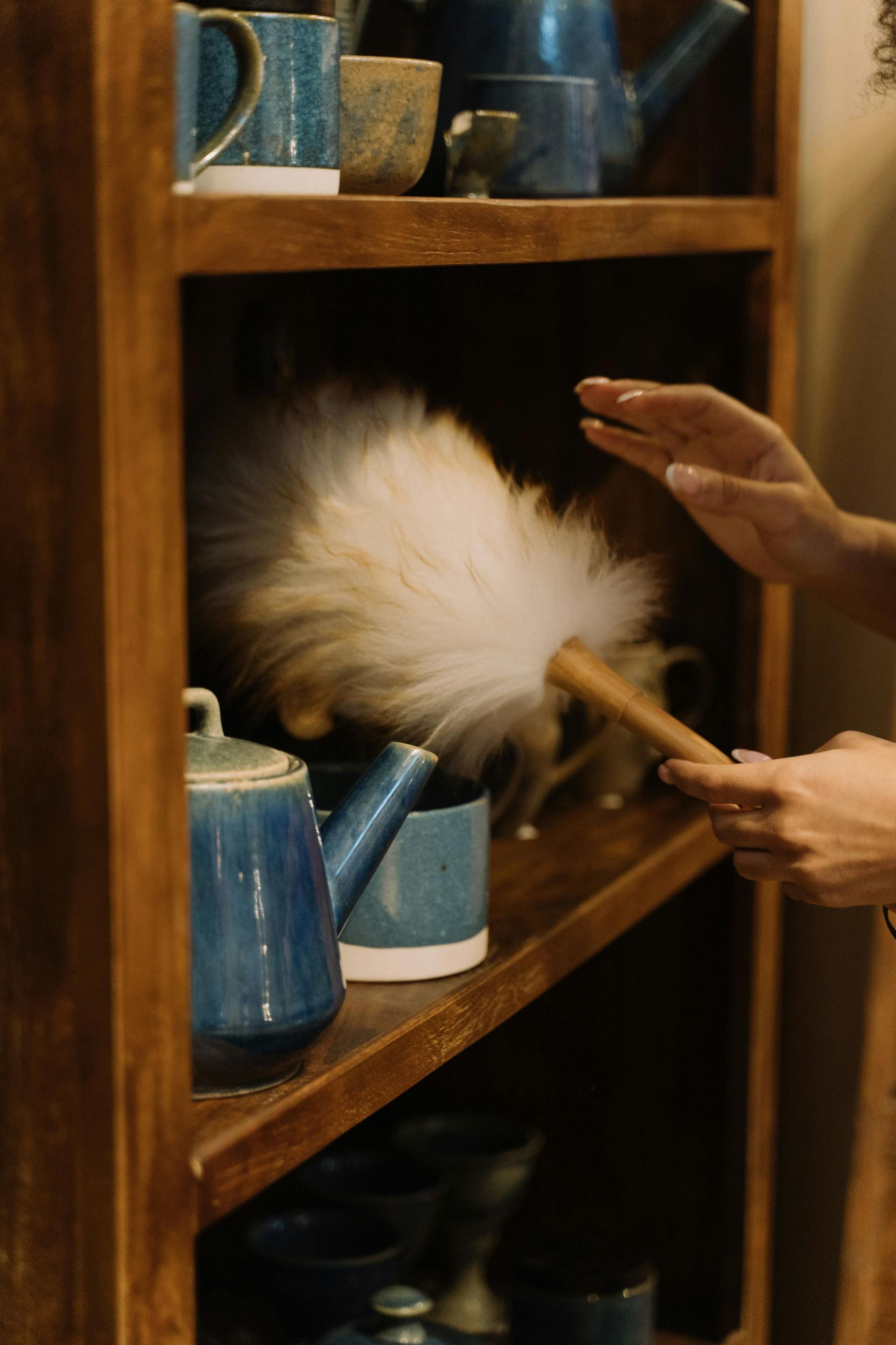 Hand dusting a wooden shelf with ceramic mugs and decor items