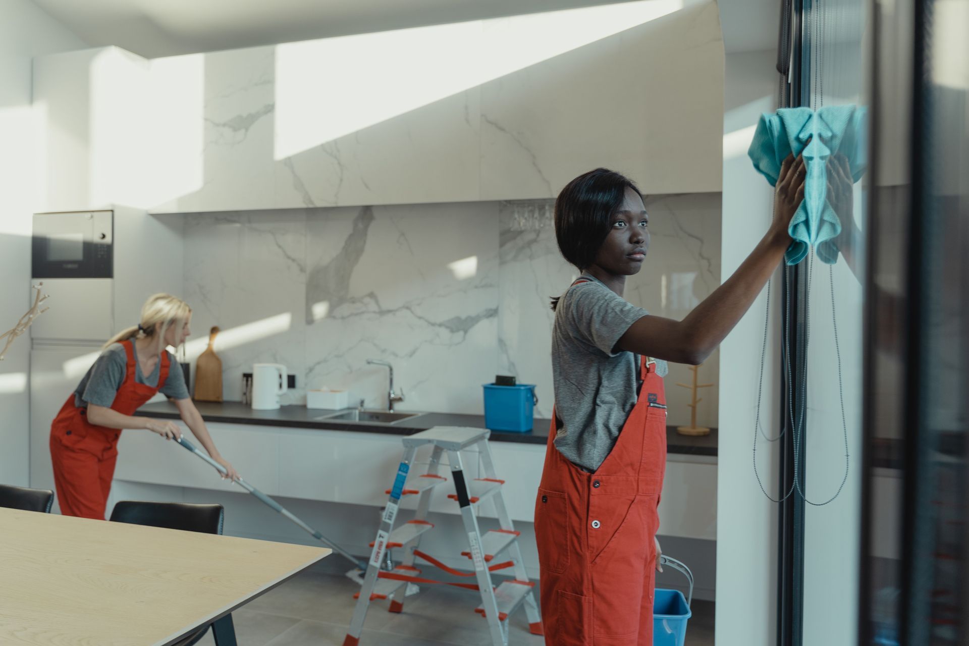 Two people cleaning a bright kitchen, one wiping a glass wall and one mopping the floor.