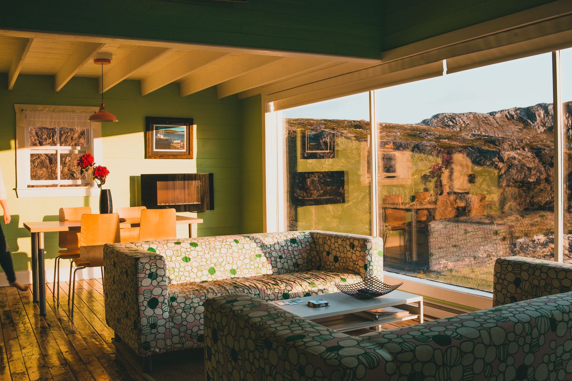 Sunlit living room with patterned sofa, dining table, and large windows overlooking a rocky landscape