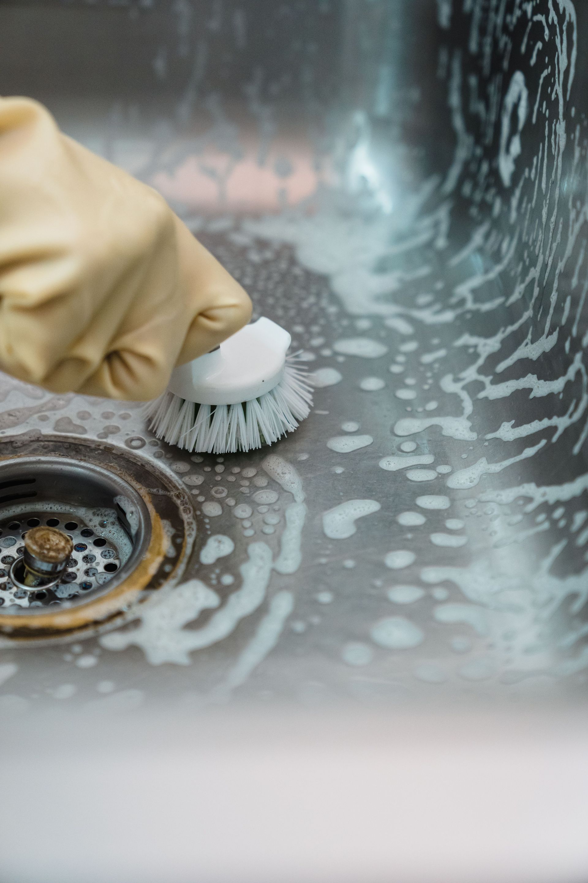 Gloved hand scrubbing a stainless steel sink with soap suds near the drain.