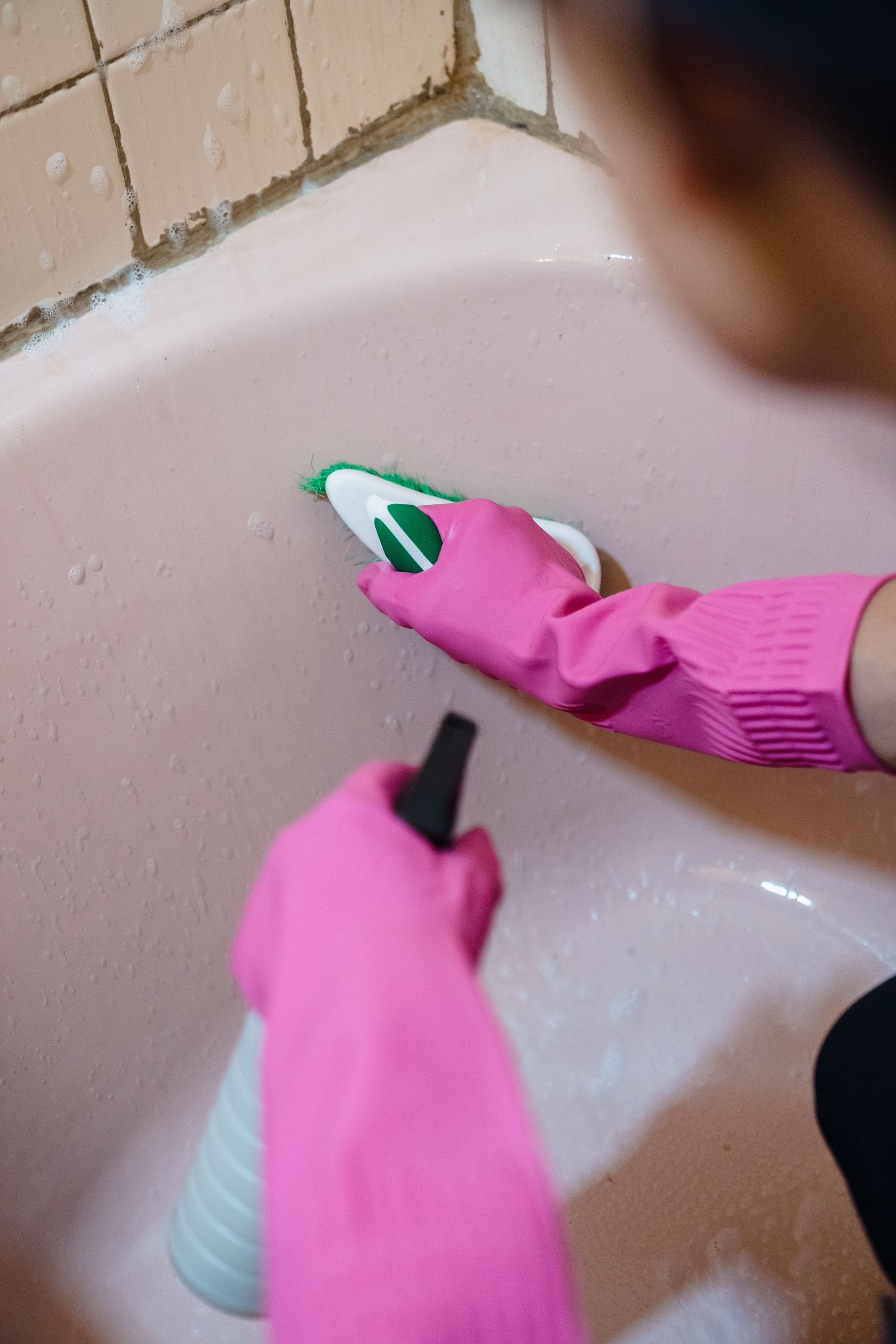 Person in pink gloves cleaning a bathtub with a spray bottle and sponge.