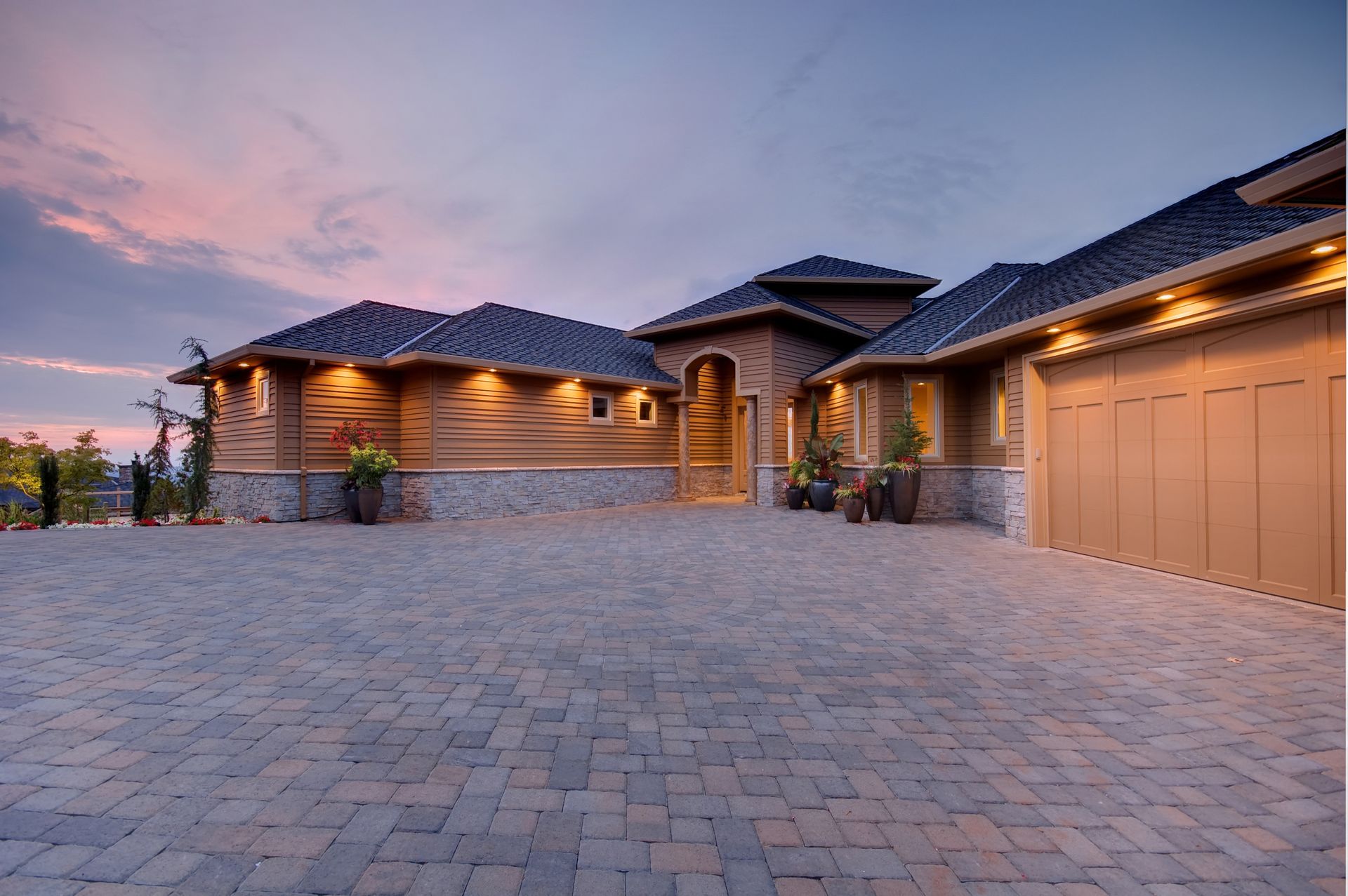 Large house with a brick driveway and tan garage under a twilight sky.
