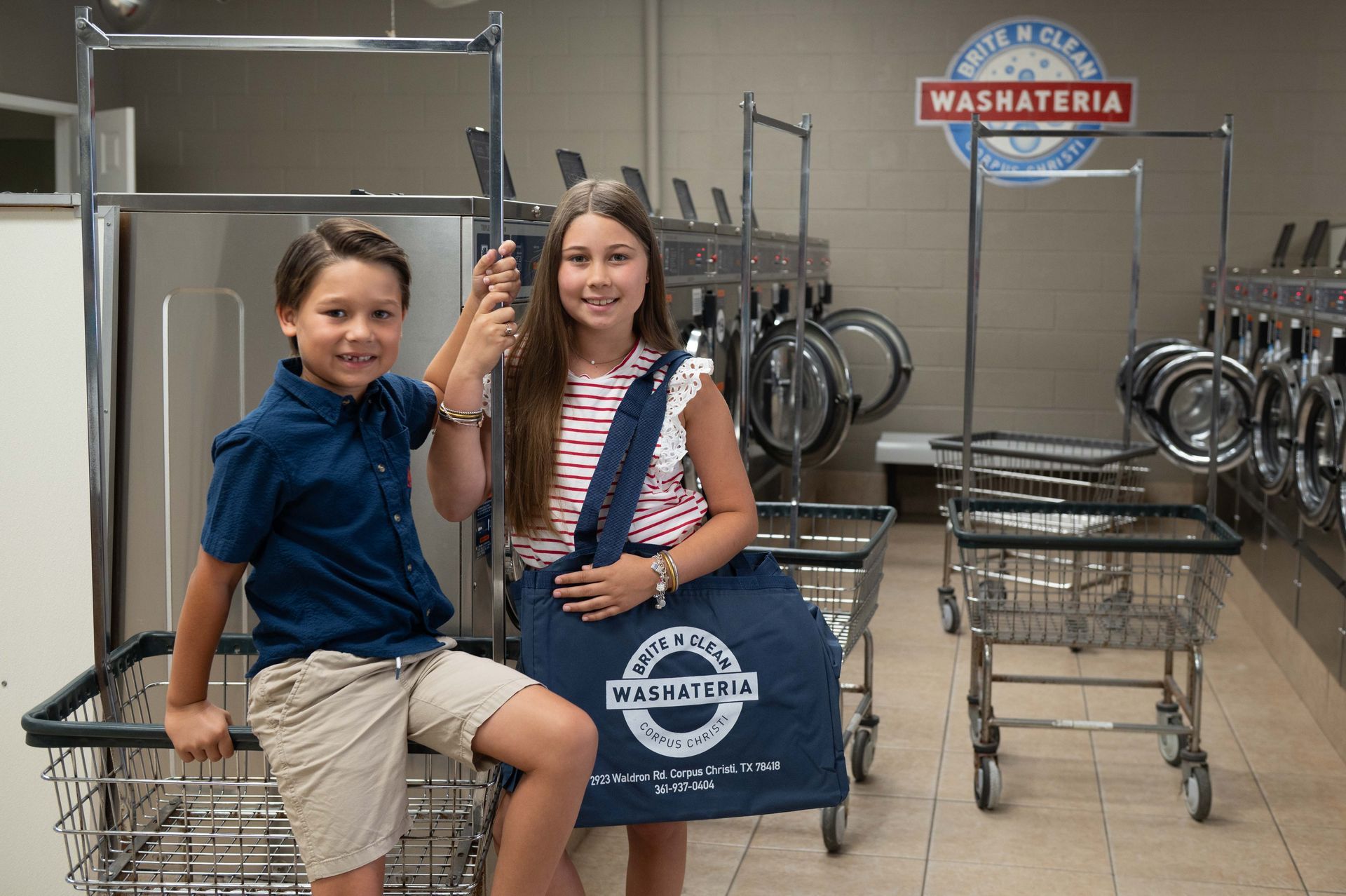 Two smiling kids at a laundromat; boy in blue shirt sits in a cart, girl holds a tote bag.