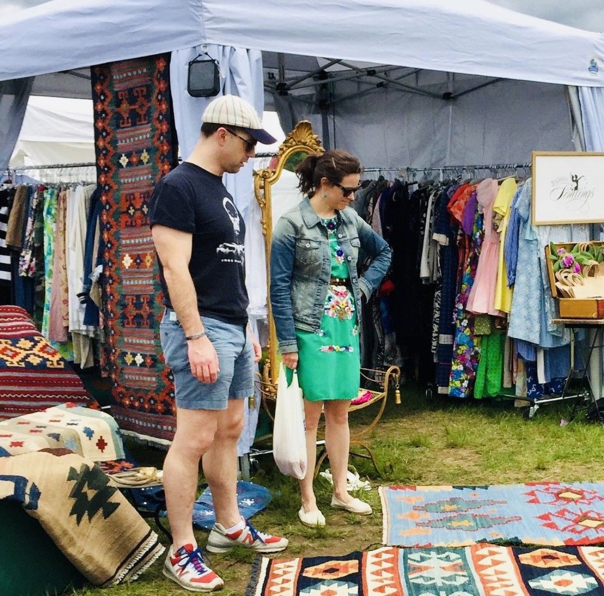 Two people browsing colorful clothes and rugs under a white market tent outdoors