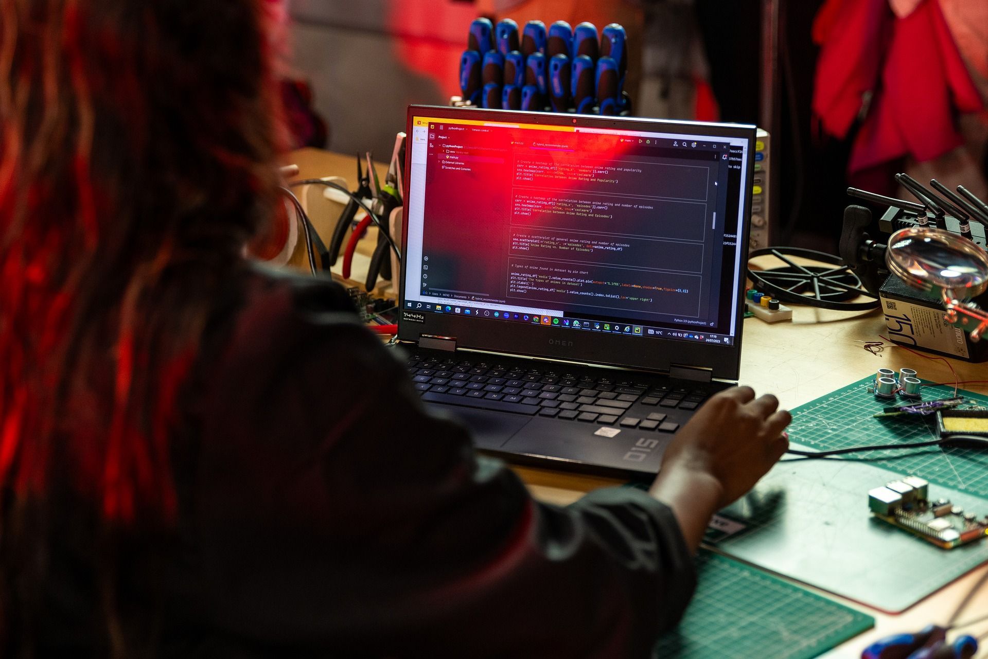 Person working on a laptop with a red-lit screen, surrounded by electronic components on a desk.