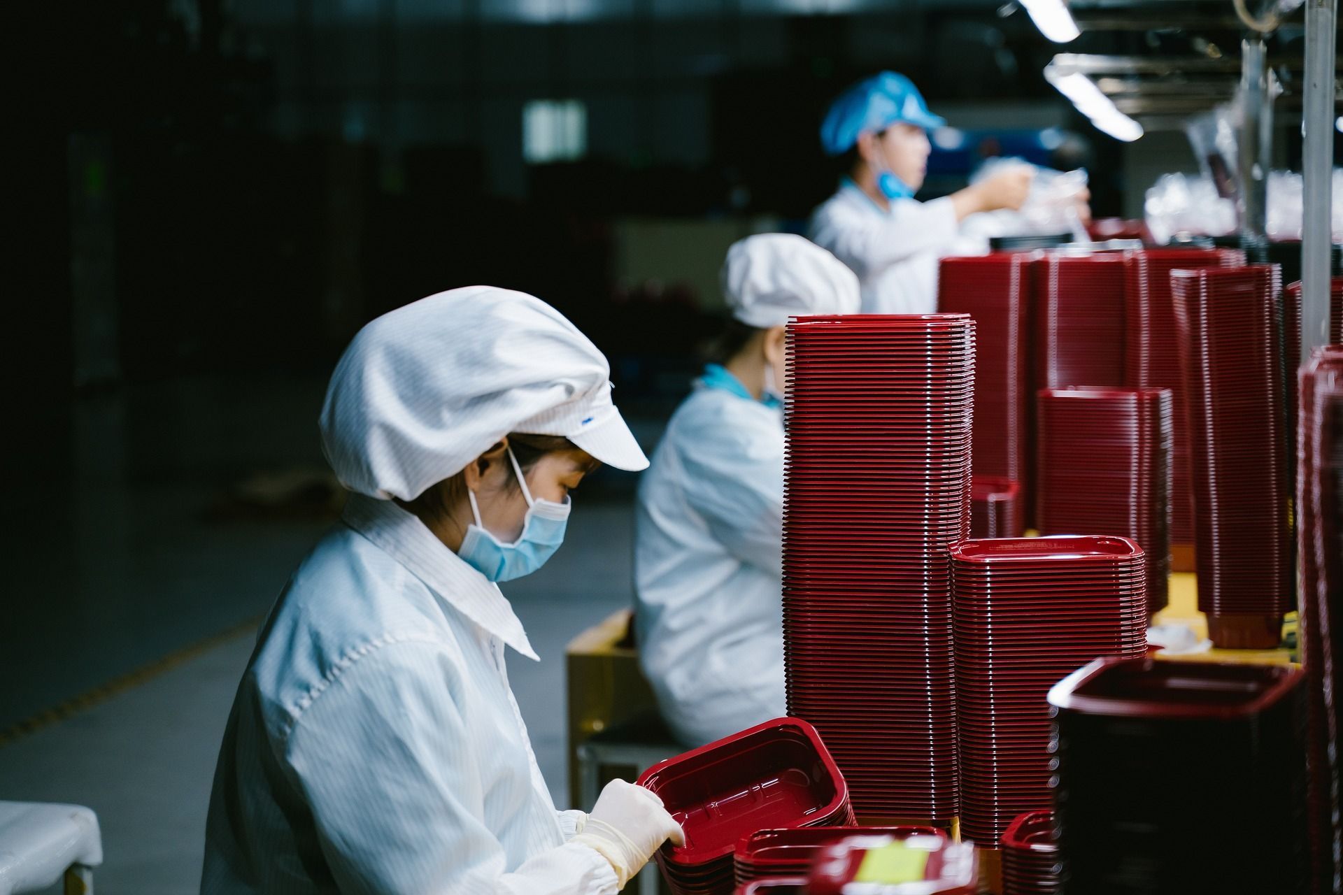 Factory workers in white coats and masks assemble red plastic containers.