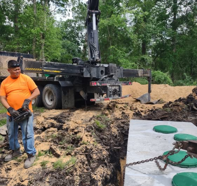 Man in orange shirt operating crane to install septic tank. Earthmoving equipment and forested background.