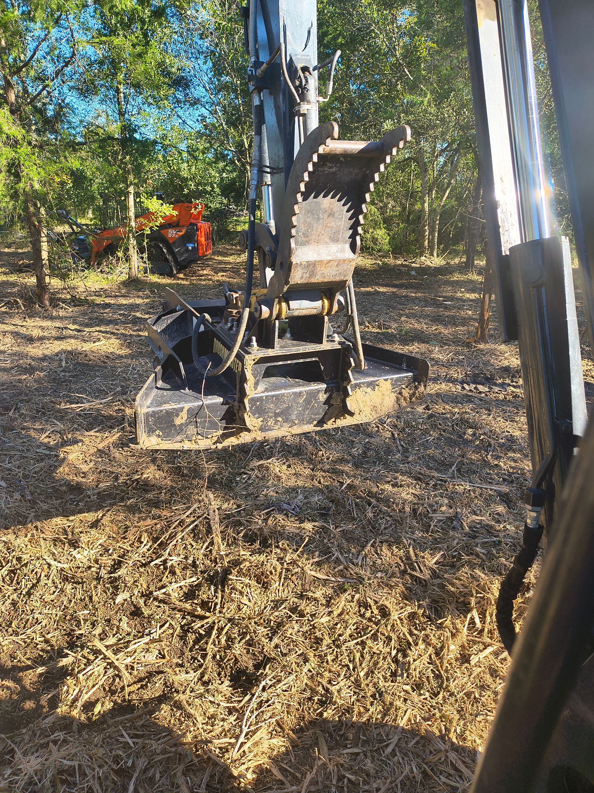 Excavator with wood chipper attachment in a wooded area, surrounded by wood chips.