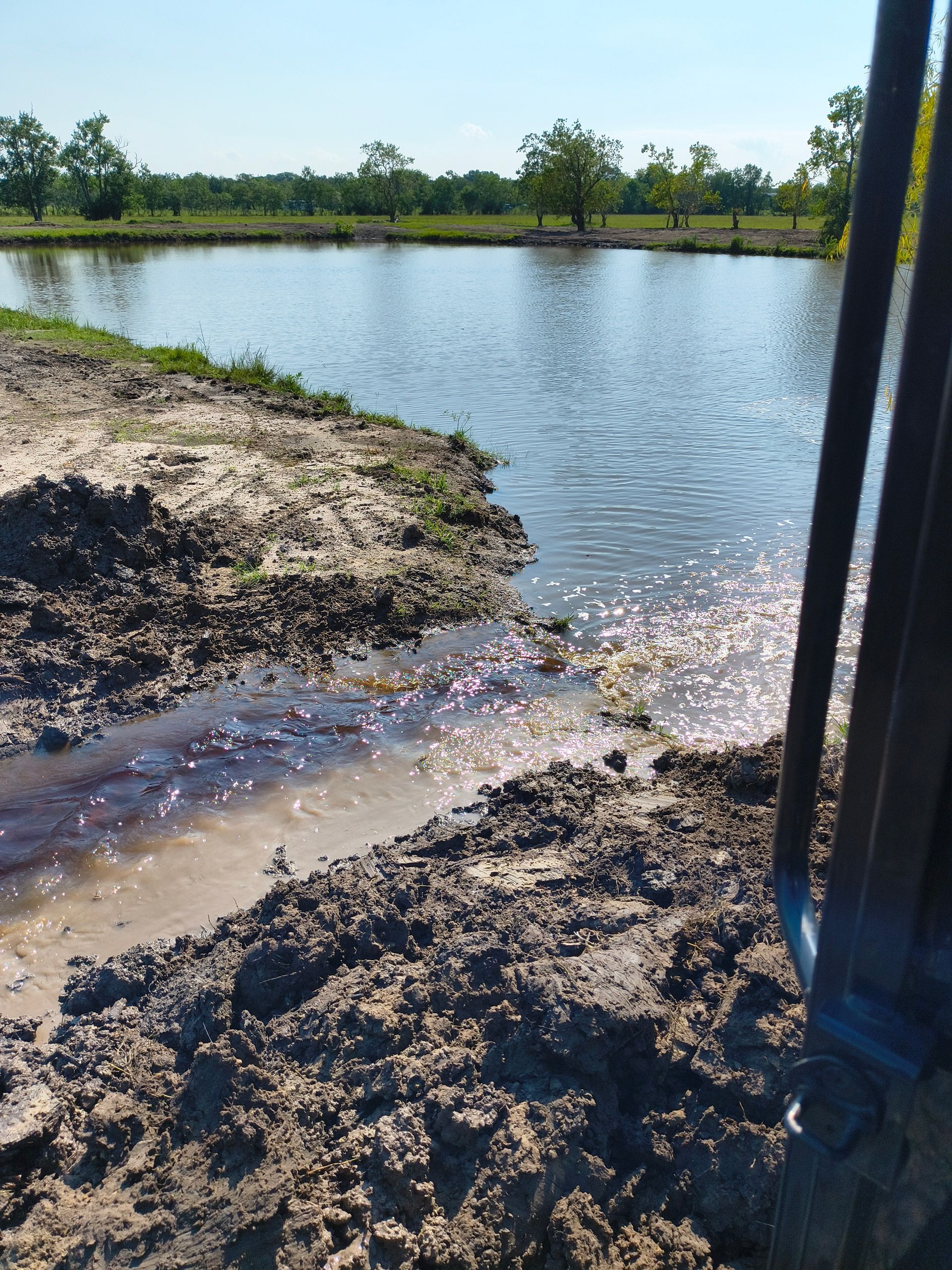 Muddy shoreline of a pond, with water, trees in the background, and a metal structure on the right side.