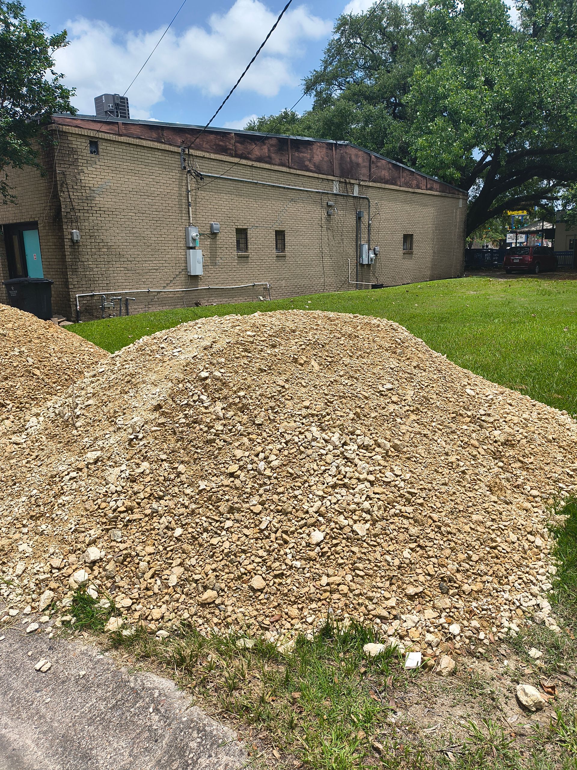 Large pile of beige gravel on a grassy area, in front of a tan brick building.