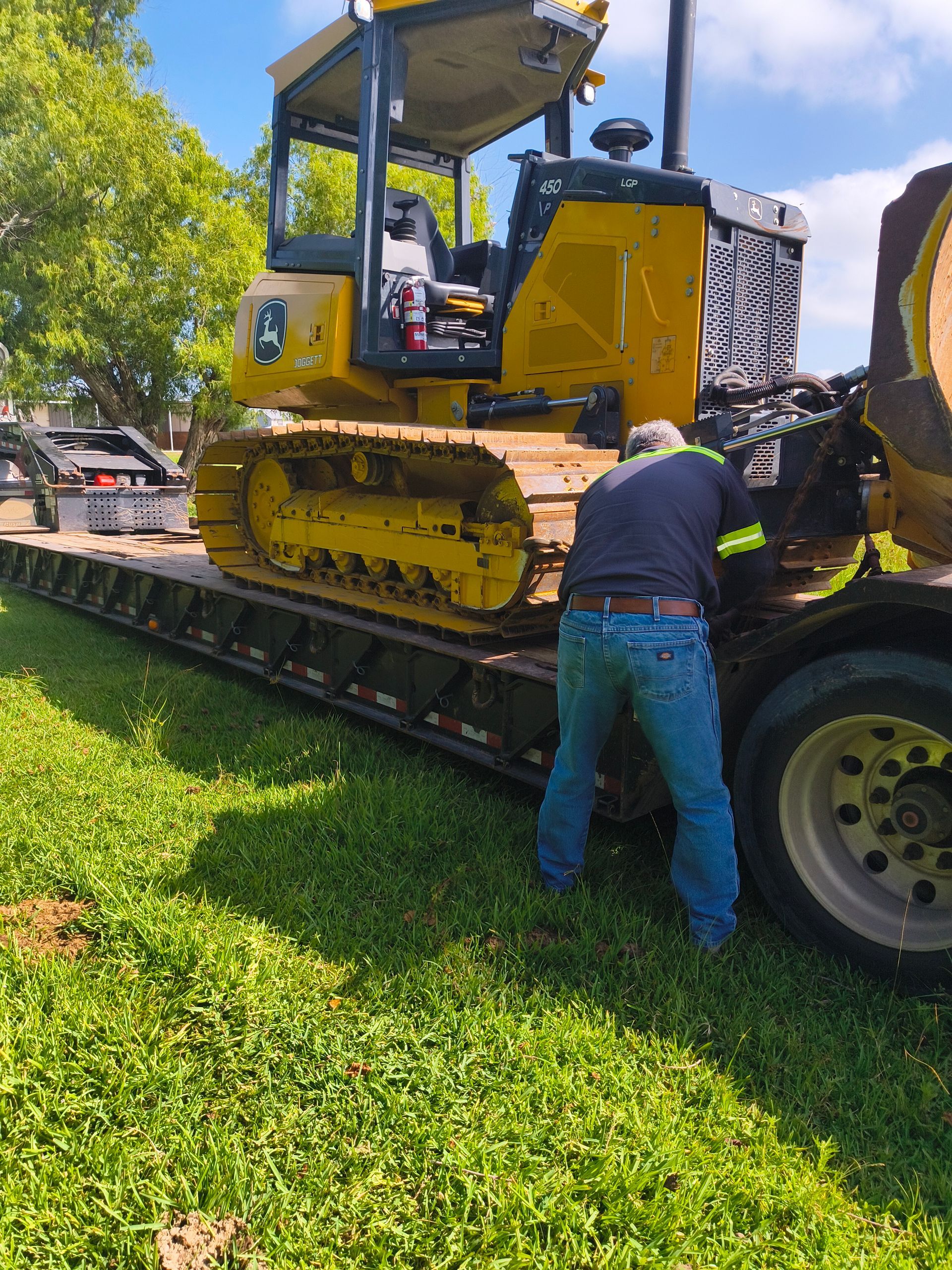 A person in jeans securing a yellow bulldozer on a flatbed trailer, outdoors on grass.