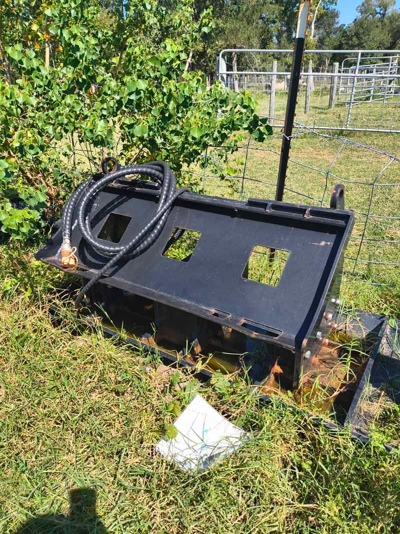 Black skid steer attachment with hose on grassy ground near foliage and a fence.