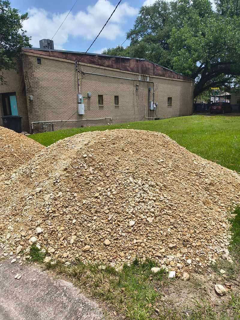 Pile of tan rocks on grass next to a tan brick building with electrical boxes and a green door.