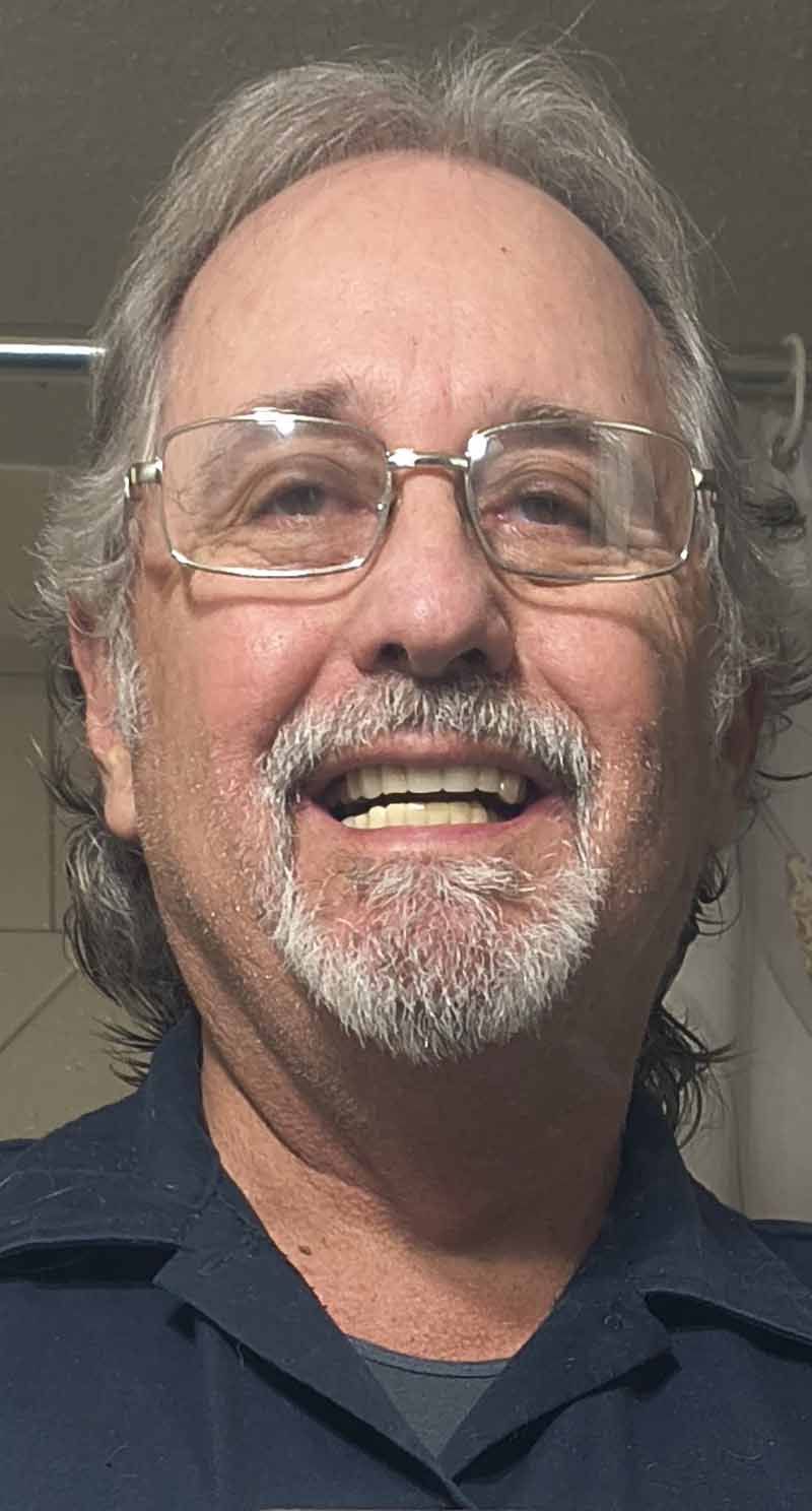 Man with gray hair and beard smiles, wearing glasses and a blue shirt.