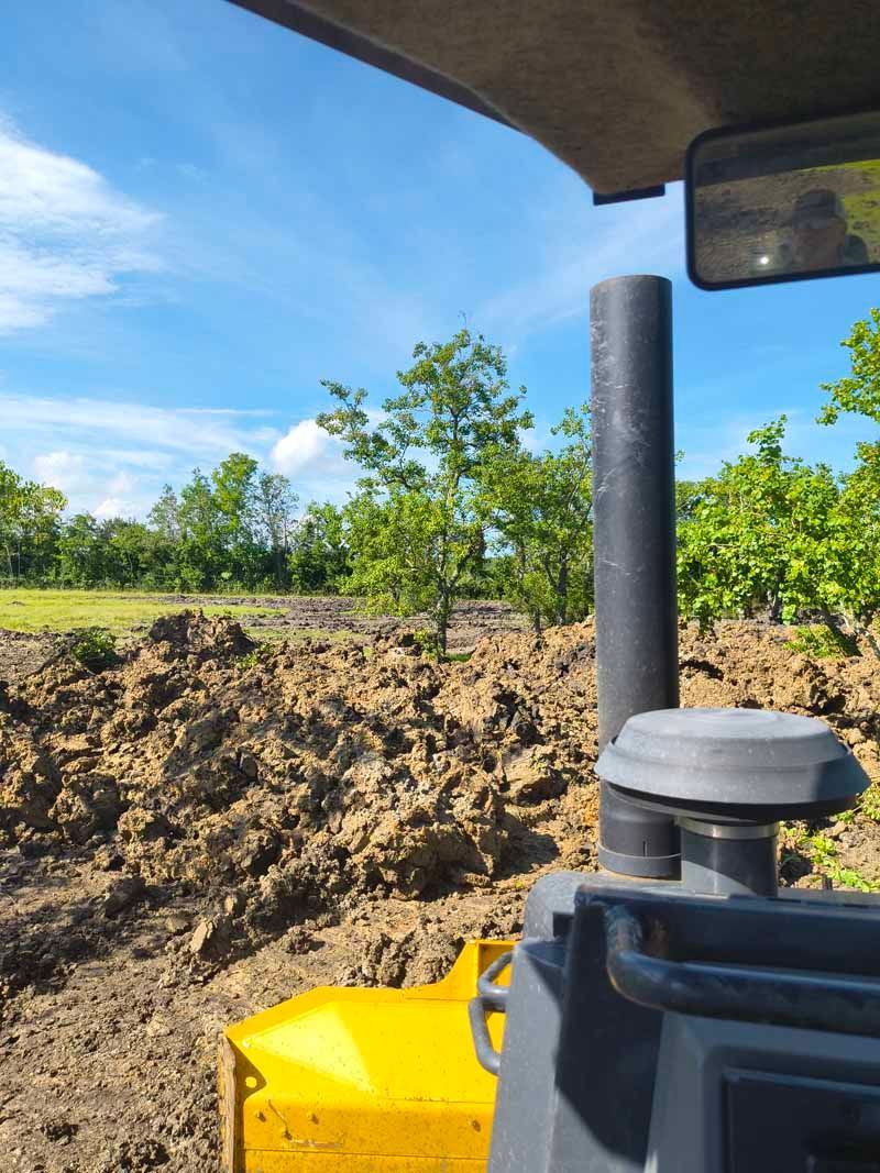 View from a bulldozer cab, showing a muddy construction site under a blue sky with trees.