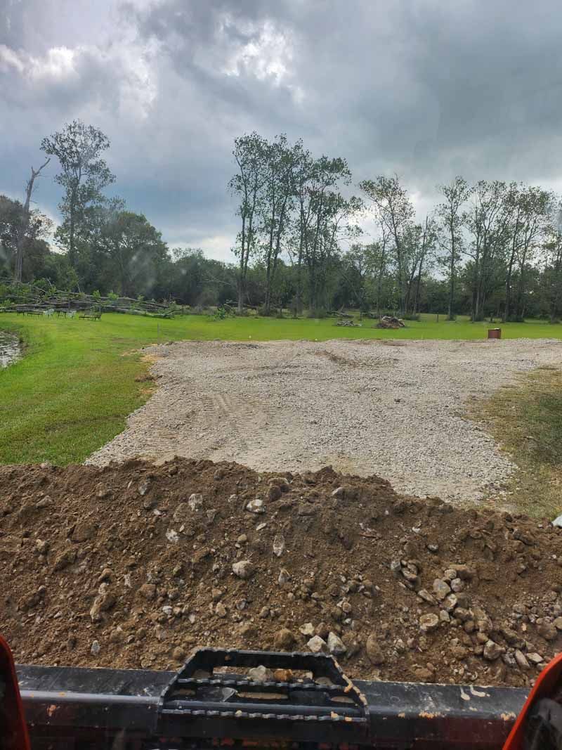 A pile of dirt in front of a gravel pathway, with a green field and trees in the background under a cloudy sky.