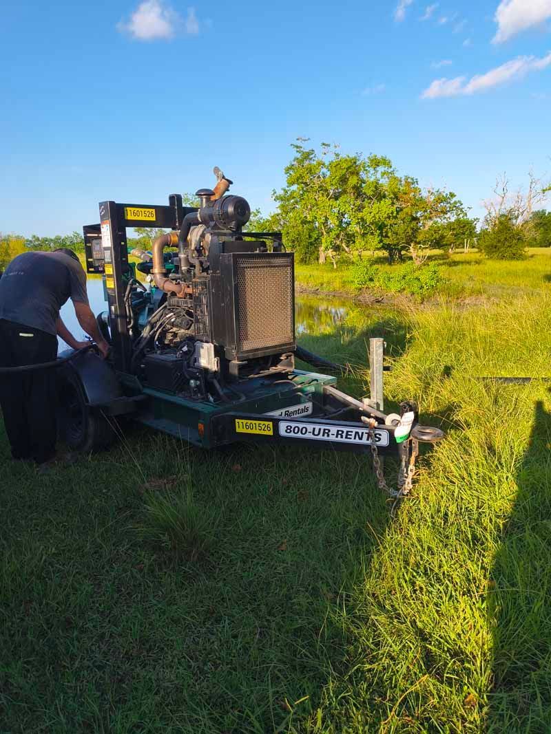Person working on a water pump on a trailer in a grassy field, under a blue sky.