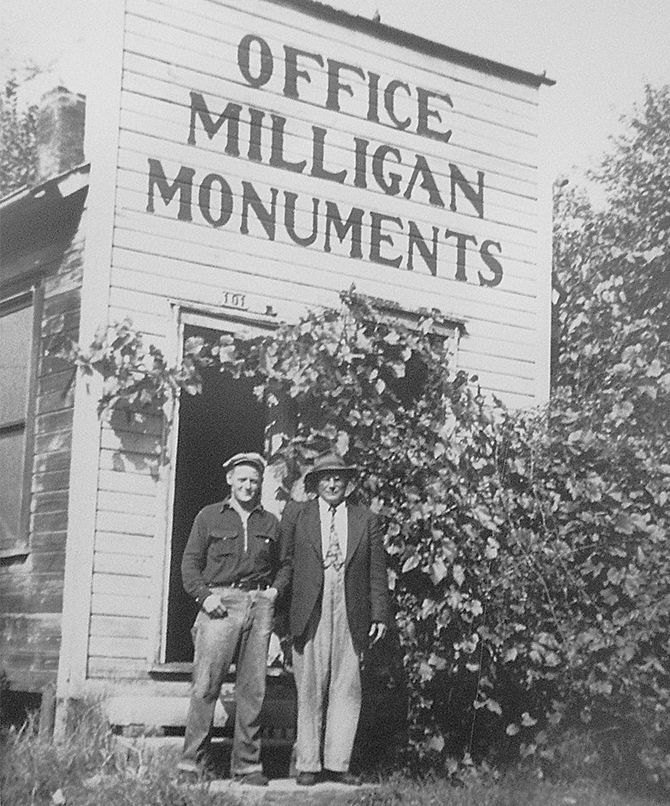 Two men stand outside Milligan Monuments office. Sign above door. Old wooden building, overgrown with plants.