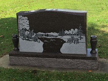Black granite headstone with etched landscape scene, set in a grassy area.
