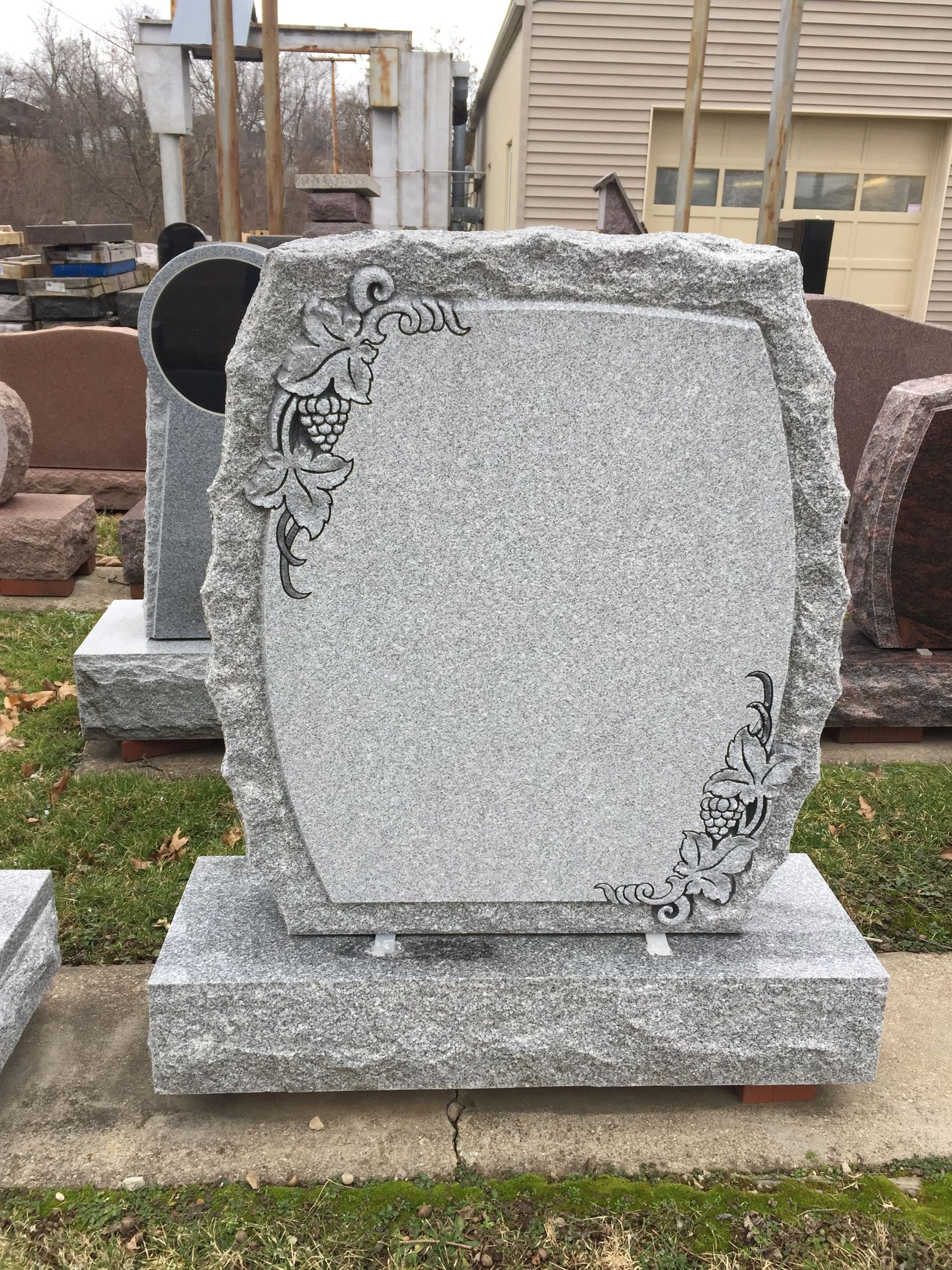 Gray granite headstone with etched floral design and a rough-hewn border, sitting on a rectangular base.
