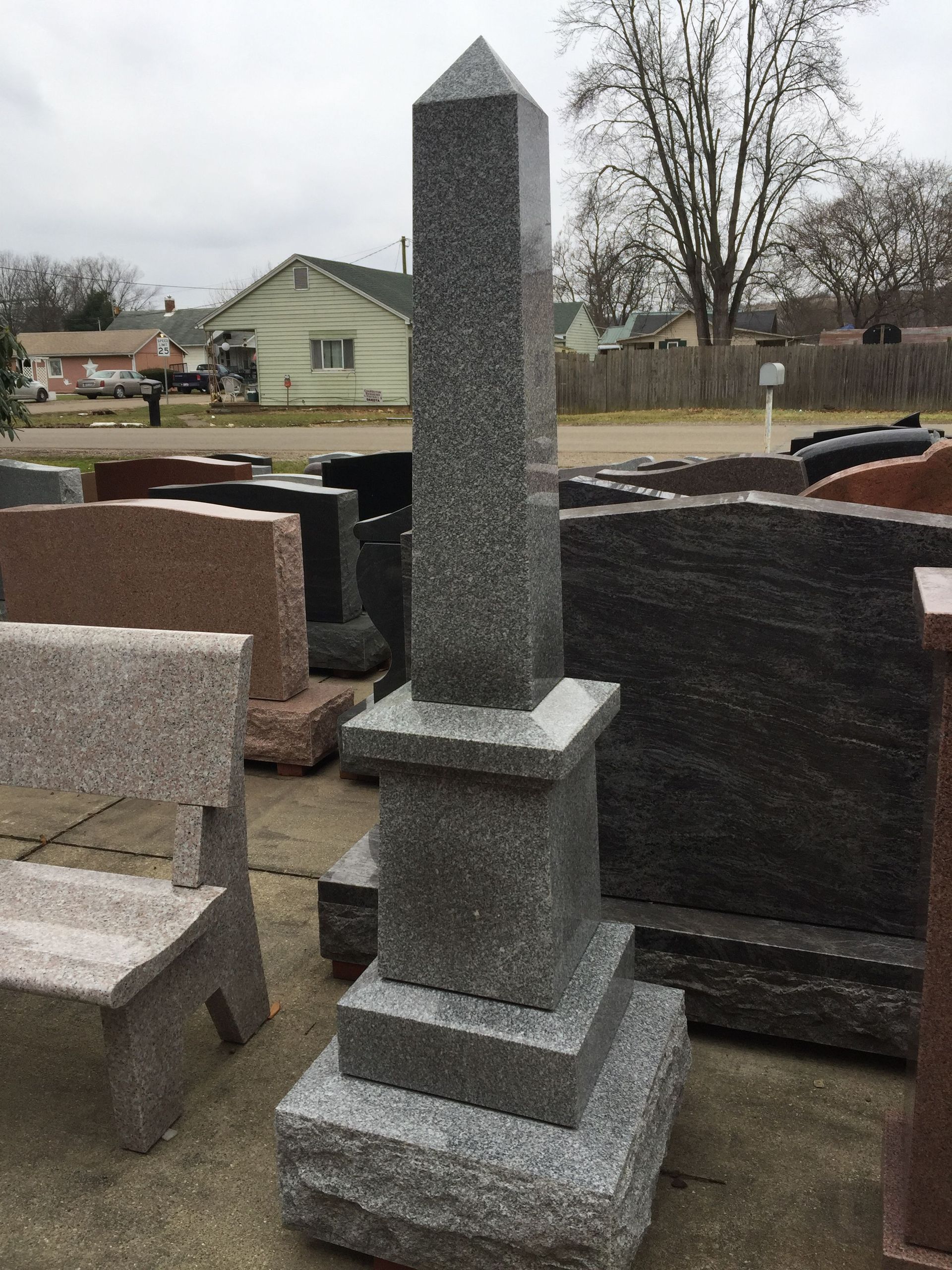 Tall gray granite obelisk tombstone displayed outside, other headstones in the background.