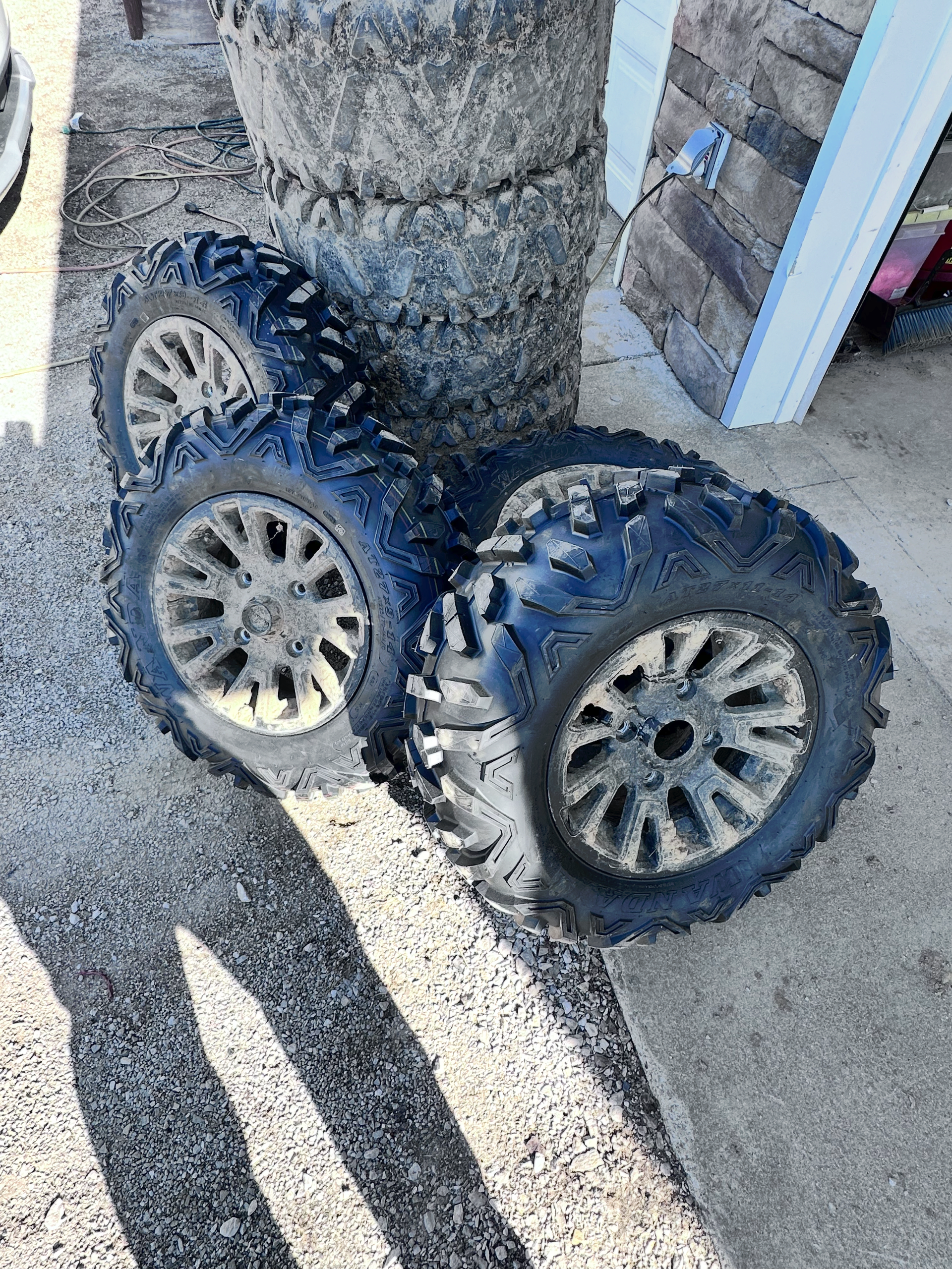 Four off-road tires with dirty rims stacked on gravel, next to a stack of more tires and a wooden wall. | Midwest Tire & Service LLC