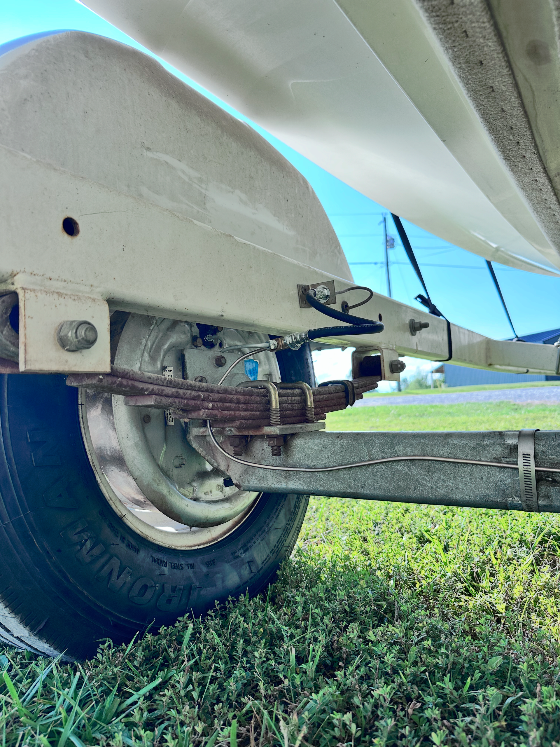 Boat trailer wheel and suspension on grass, close-up. White hull, black tire, rusty leaf springs. | Midwest Tire & Service LLC