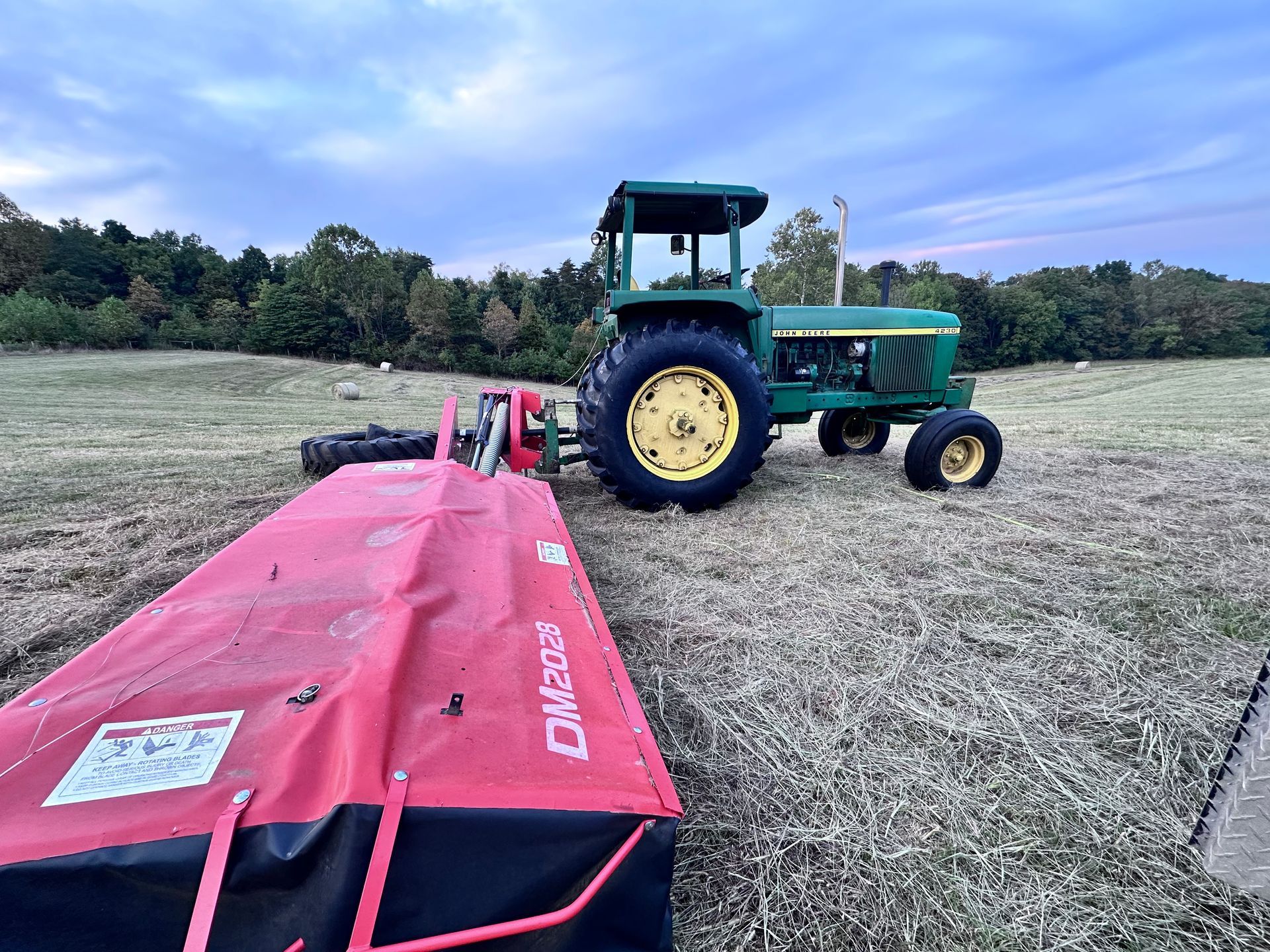 Green John Deere tractor with a red mower in a field under a cloudy sky. | Midwest Tire & Service LLC