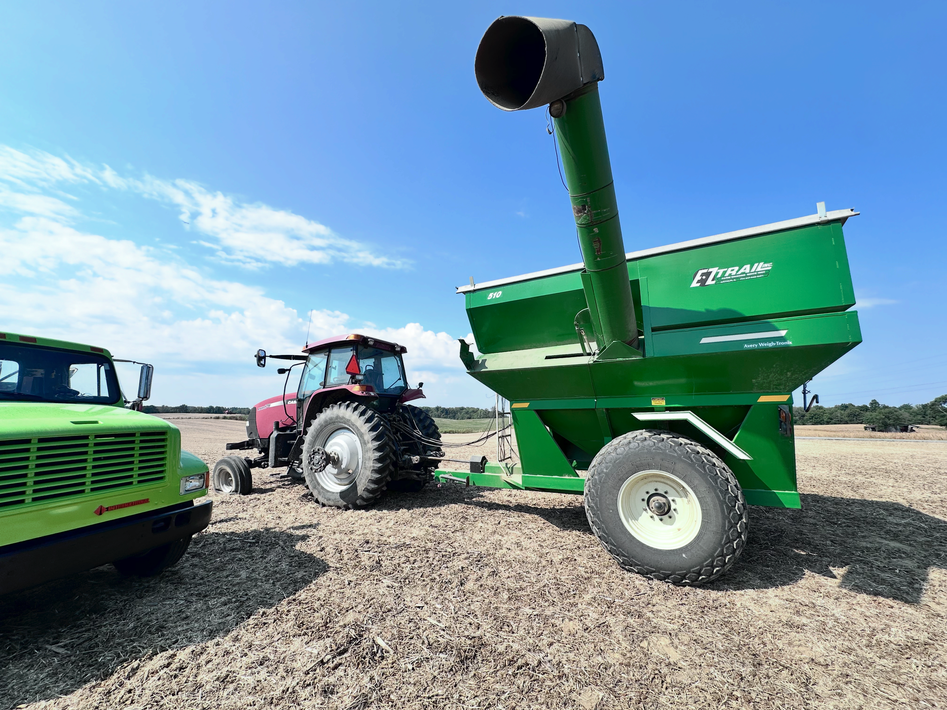 Green grain cart being pulled by a tractor in a field under a blue sky. | Midwest Tire & Service LLC