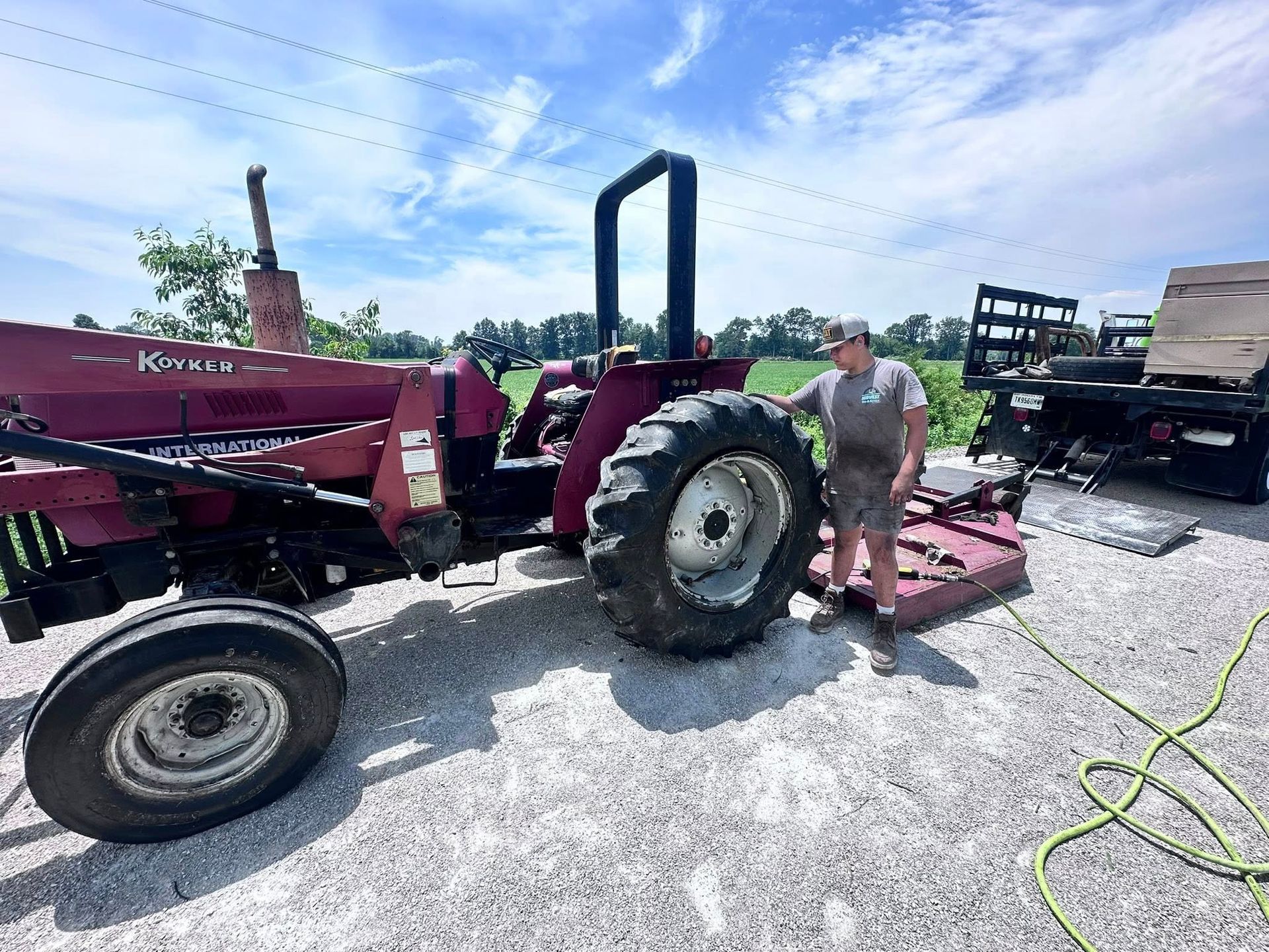 Man Standing Next to a Red Tractor on a Gravel Driveway, Under a Bright Blue Sky | Midwest Tire & Service LLC