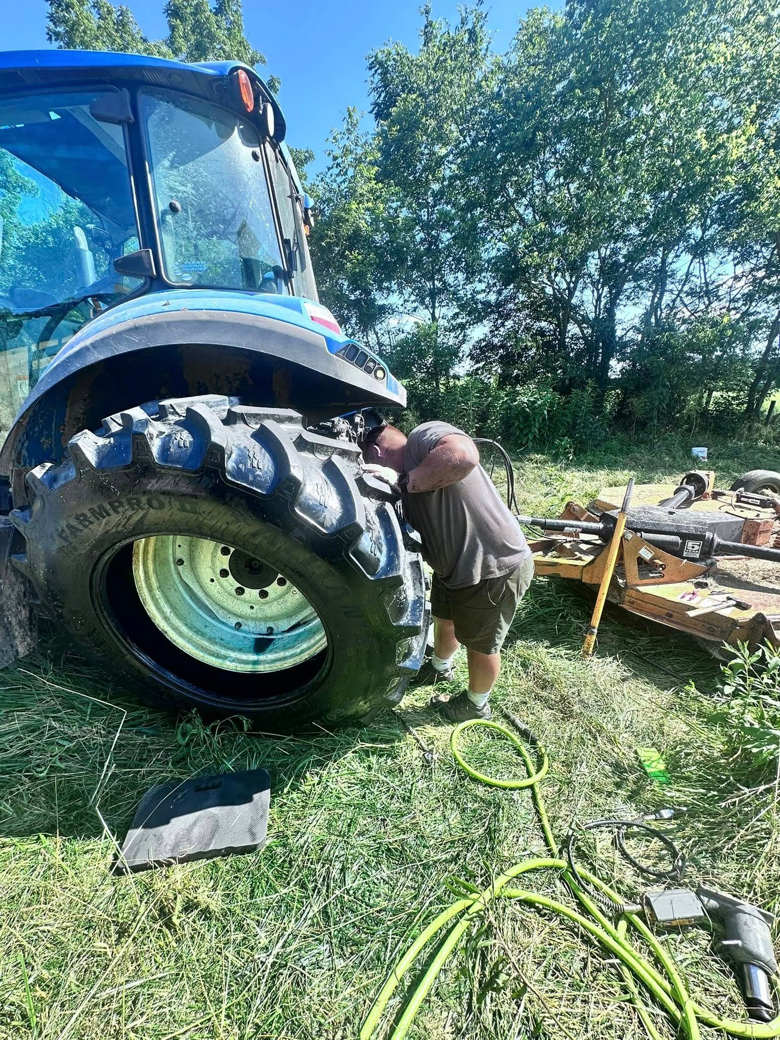 Man Working on a Tractor Tire Outdoors. Blue Tractor, Green Grass, Sunny Day | Midwest Tire & Service LLC