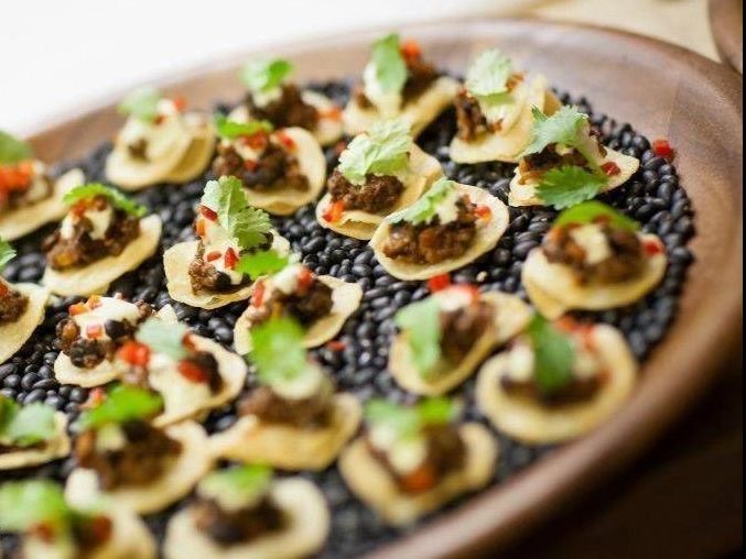 Appetizers on tortilla chips with toppings and cilantro, displayed on black beans in a wooden dish.