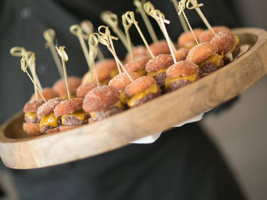 A server holding a wooden tray of mini cheeseburger sliders, secured with decorative skewers.